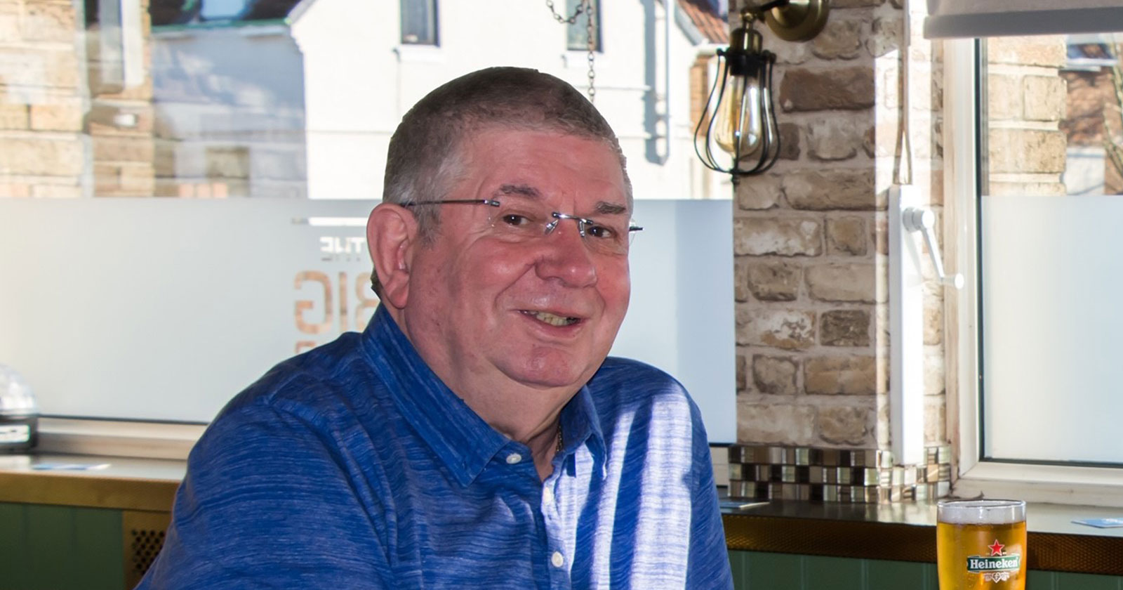 A man wearing glasses and a blue shirt sits at a table indoors with a glass of Heineken beer in front of him. There is a window and a brick wall in the background.