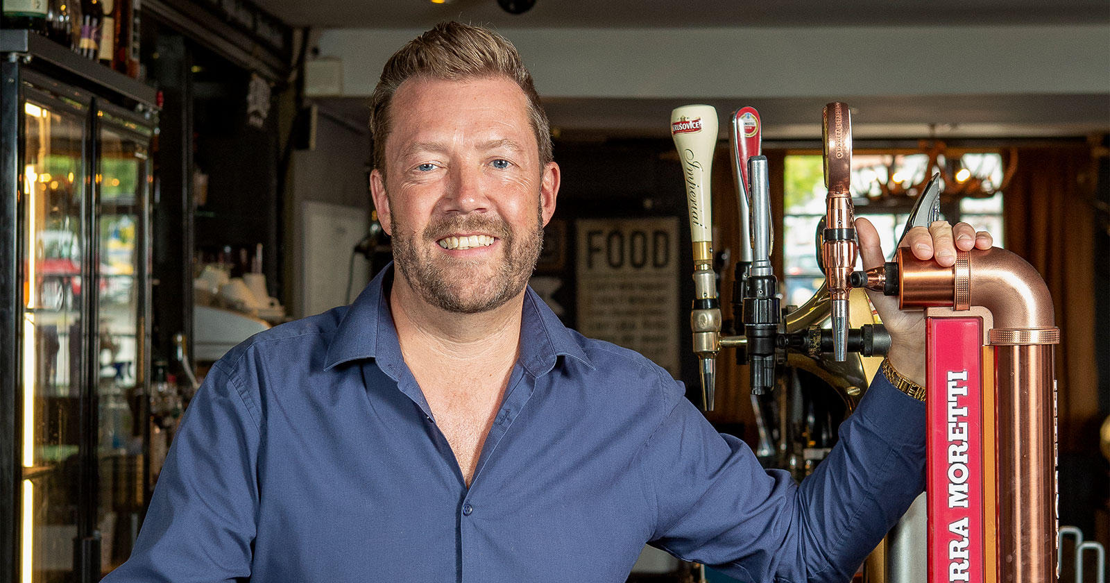 Man in blue shirt smiling and standing next to beer taps in a bar.