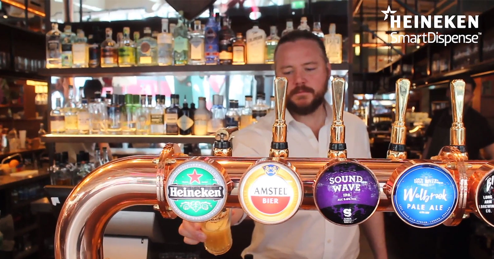 A bartender is pouring a beer from a tap at a bar. The taps include Heineken, Amstel Bier, Sound Wave IPA, and Wolf Rock Pale Ale. Shelves behind display various bottles of alcohol.