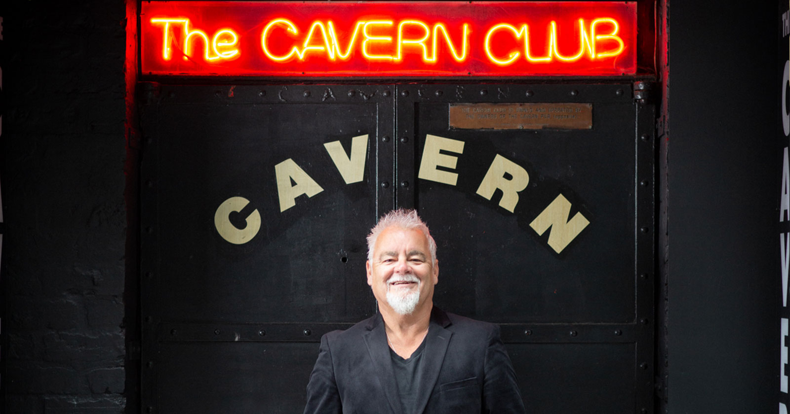 A man with white hair and beard stands smiling in front of The Cavern Club entrance, with a black door and red neon sign above.