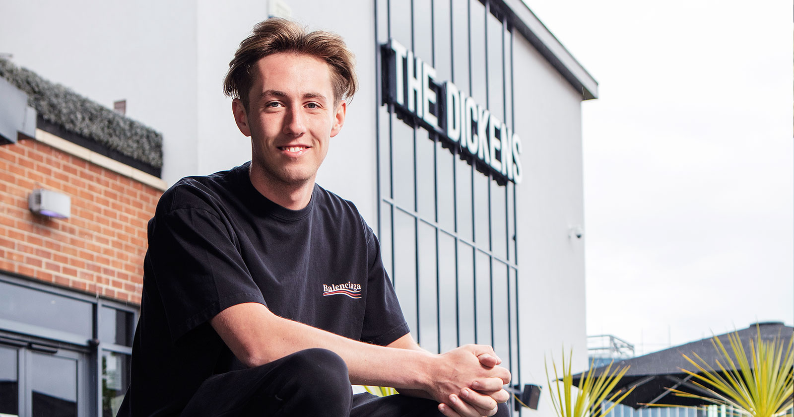 A person in a black shirt sits outside a building with a sign that reads "THE DICKENS.