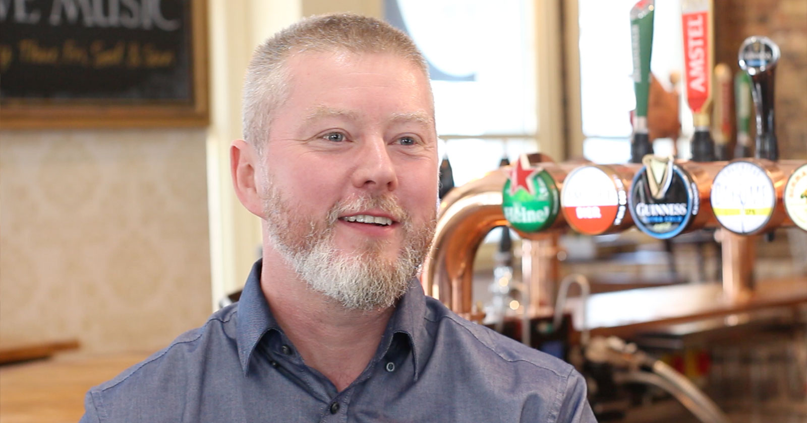 A man with a gray beard and short hair smiles while sitting in a bar. Various beer taps are visible in the background.