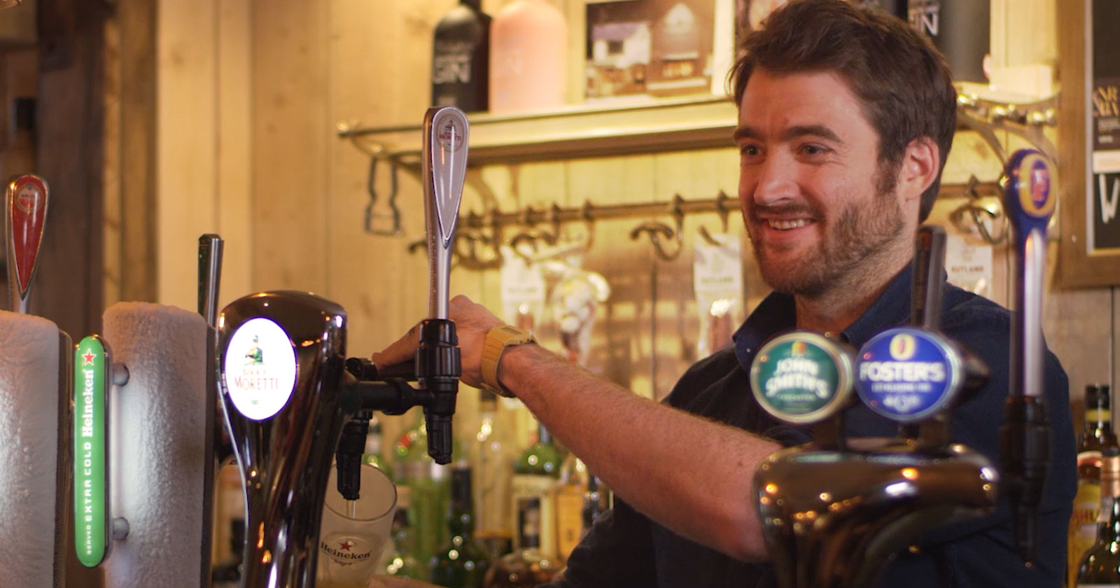A man with a beard smiles while serving a drink behind a bar, with various beer taps and bottles visible in the background.