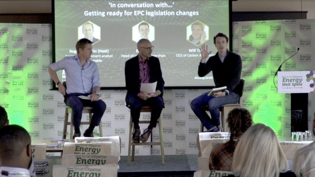 Three men are sitting on stage during a discussion panel at the Energy Well Spent event about EPC legislation changes. One man on the right is speaking and gesturing, the two others are listening.