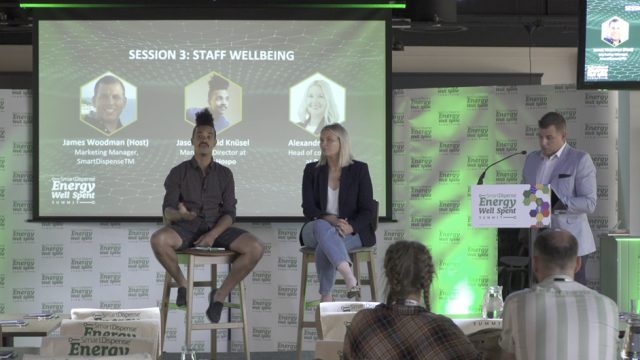 Three speakers and a moderator are on a stage for a "Staff Wellbeing" session at an Energy conference. Two speakers are seated, one is standing at a podium. Conference banners are in the background.