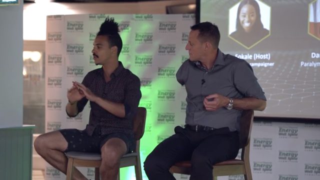 Two individuals, one with a unique hairstyle and wearing casual clothing, and the other wearing a button-up shirt, are seated on chairs, engaged in a conversation on a stage with "Energy" branding.