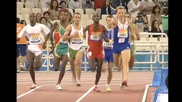 Runners compete in a track race, with spectators watching from the stands.