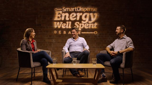 Three people are seated in armchairs on a stage with a sign that reads "SmartDispense Energy Well Spent Debate" on the brick wall behind them. They appear to be engaged in a discussion.