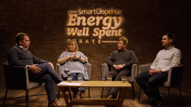 Four people are sitting in chairs in a semi-circle having a discussion. The background wall displays the text "SmartDispense Energy Well Spent Debate.
