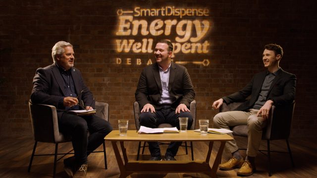 Three men sitting on a stage in a discussion setup with the backdrop displaying "SmartDispense Energy Well Spent Debate". They are engaged in conversation, with water glasses and notes on the table.