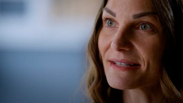 Close-up of a woman with long brown hair, slightly smiling and looking to the side. She has a neutral expression and the background is blurred.