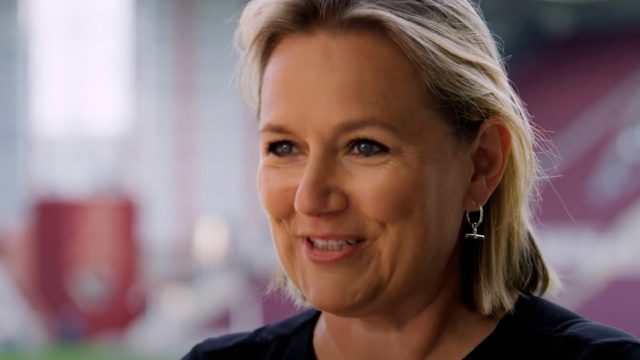 A person with short blonde hair, wearing a black top and cross earrings, is smiling against a blurred background.