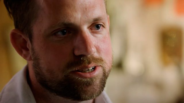A man with short hair and a beard is speaking, with a neutral expression and a blurred background.