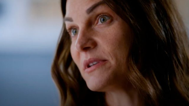 Close-up of a woman with long brown hair and green eyes, looking slightly to the side. She has a neutral expression on her face. The background is blurred.