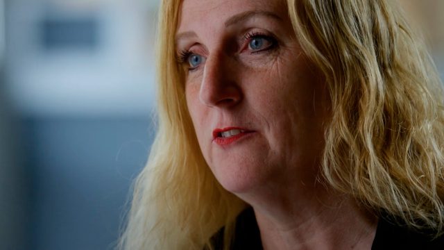 Close-up of a woman with long blonde hair and blue eyes, wearing a black top, looking slightly off-camera in a lit indoor setting.