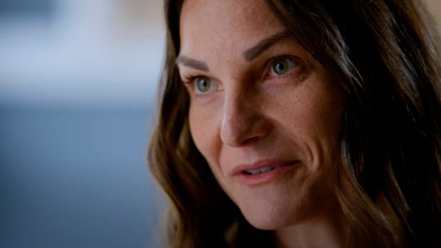 Close-up of a woman with long brown hair, green eyes, and a neutral expression, looking slightly to the side. The background is softly blurred.