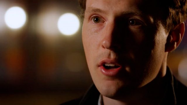 Close-up of a person with short hair speaking, with unfocused lights in the background.