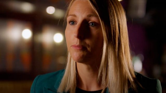 A woman with blonde hair and a teal jacket looks forward with a serious expression. Blurred lights are in the background.