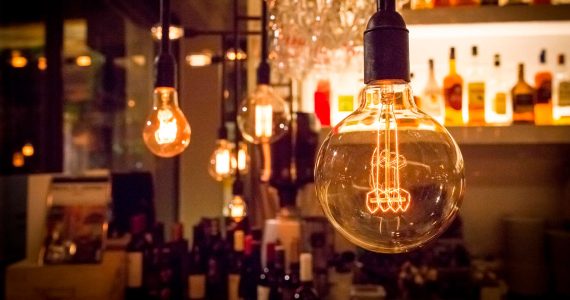 Close-up of hanging Edison-style light bulbs with a warm glow, illuminating a bar area stocked with various bottles.