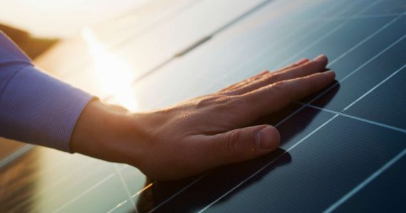 A hand touching the surface of a solar panel in the sunlight.