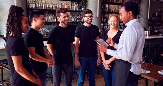 Five people in black shirts stand in a semicircle, listening to a person in a white shirt holding a tablet, in a bar setting with shelves of bottles in the background.