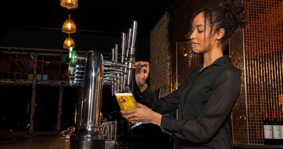 A bartender with curly hair pours a drink from a tap into a yellow cup at a dimly lit bar.