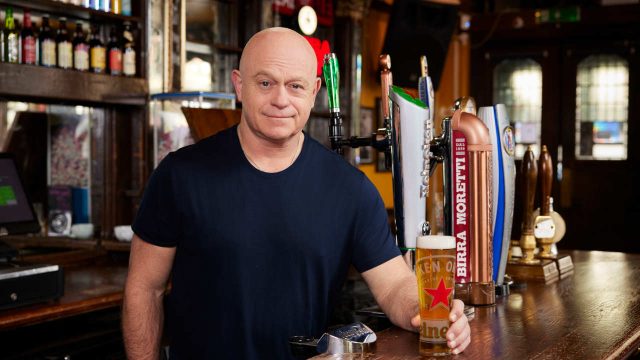 A person stands behind a bar holding a pint of beer, with beer taps and bottles visible in the background.