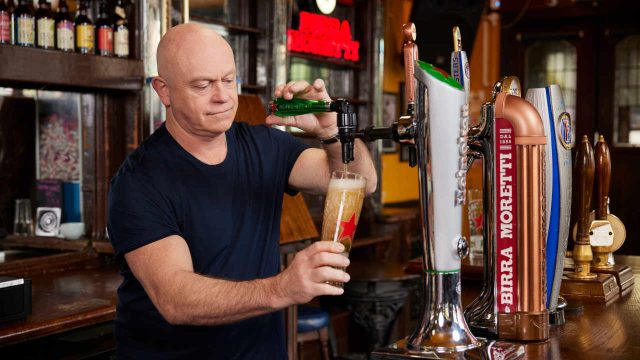 Man with a shaved head in a black t-shirt pouring beer from a tap into a glass in a pub with various beer taps and bottles visible in the background.