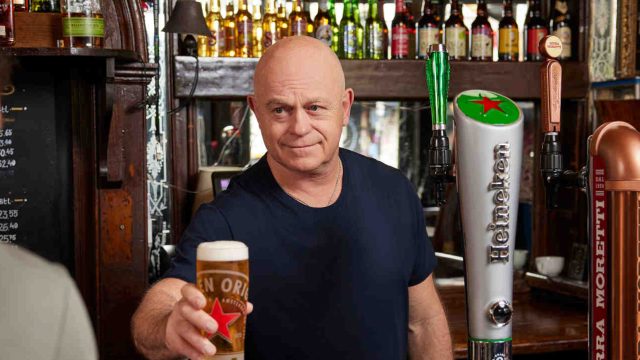 A bartender in a black shirt serves a pint of beer to a customer in a pub. Various beer taps and bottles are visible behind the bar.